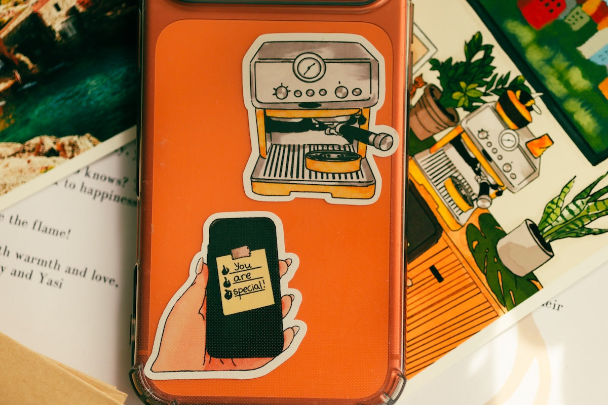 An orange phone case with two cartoon stickers, displayed on top of illustrated books. The stickers depict a coffee machine and a hand holding a phone with a 'Cafe Special' note. Detailed view of custom vinyl stationery stickers featuring a coffee machine and a post-it note attached to a phone case