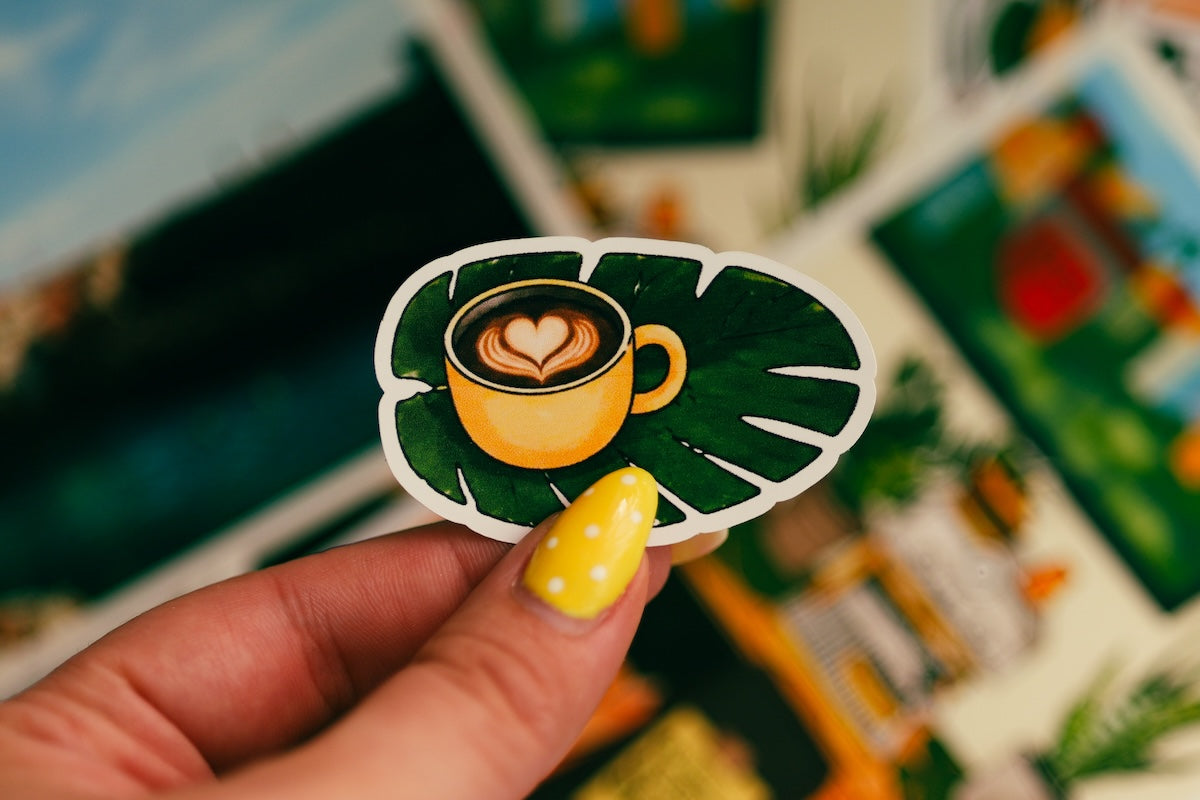 A hand with yellow nail polish holds a die-cut sticker featuring a yellow coffee cup with heart latte art on a green monstera leaf, with blurred tropical cards in the background. Close up of a hand holding a custom illustrated coffee sticker over creative aesthetic postcards