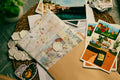 A styled flat-lay of a handmade paper craft journal with floral watercolor patterns and wax seals, surrounded by matching illustrated postcards, a kraft envelope, and decorative ceramic seals on a green patterned tablecloth.