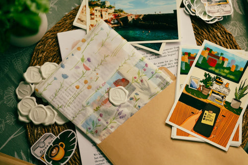 A styled flat-lay of a handmade paper craft journal with floral watercolor patterns and wax seals, surrounded by matching illustrated postcards, a kraft envelope, and decorative ceramic seals on a green patterned tablecloth.