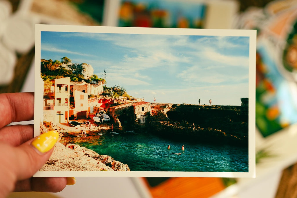 A hand with yellow nail polish holds a printed photograph or postcard showing a coastal village with blue water, rocky cliffs, and buildings under a bright sky. The background is softly blurred with other colorful items visible. A hand with yellow polka-dot nails holding a vibrant horizontal photo print of a Mediterranean seaside town with turquoise water