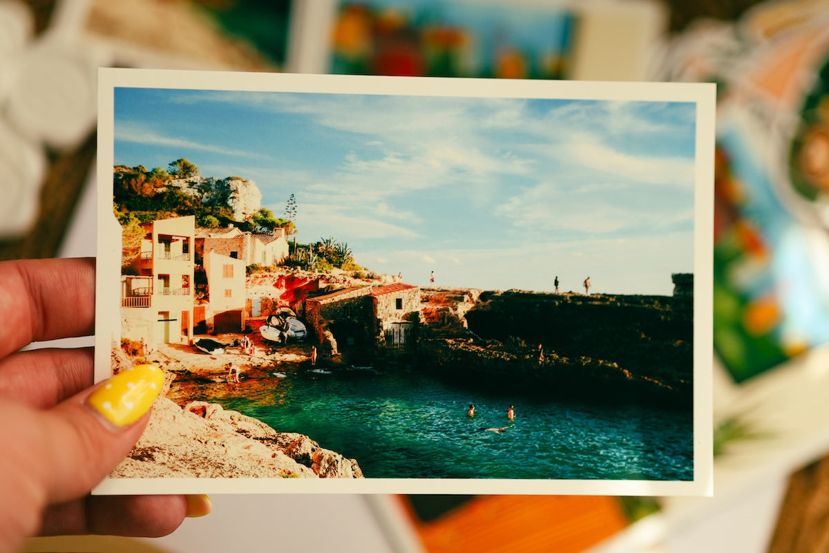 A hand with yellow nail polish holds a printed photograph or postcard showing a coastal village with blue water, rocky cliffs, and buildings under a bright sky. The background is softly blurred with other colorful items visible. A hand with yellow polka-dot nails holding a vibrant horizontal photo print of a Mediterranean seaside town with turquoise water