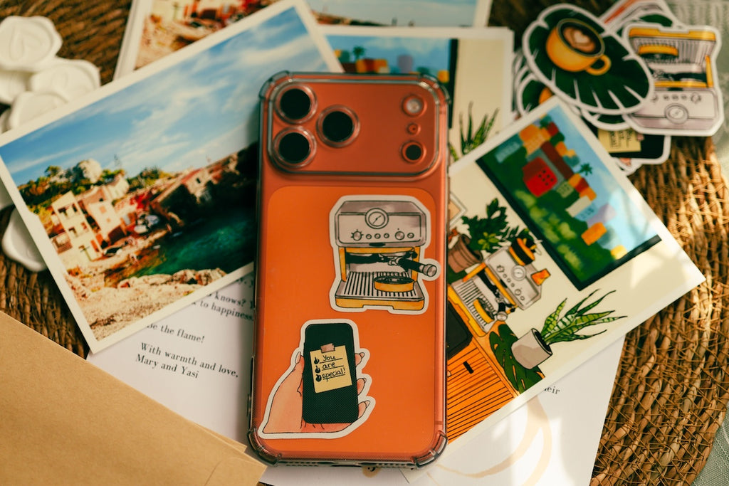 An orange phone case with retro-style stickers, shown on a phone and surrounded by colorful illustrated postcards on a woven surface. An orange smartphone decorated with custom artistic stickers, surrounded by beautiful postcards and white wax stamps