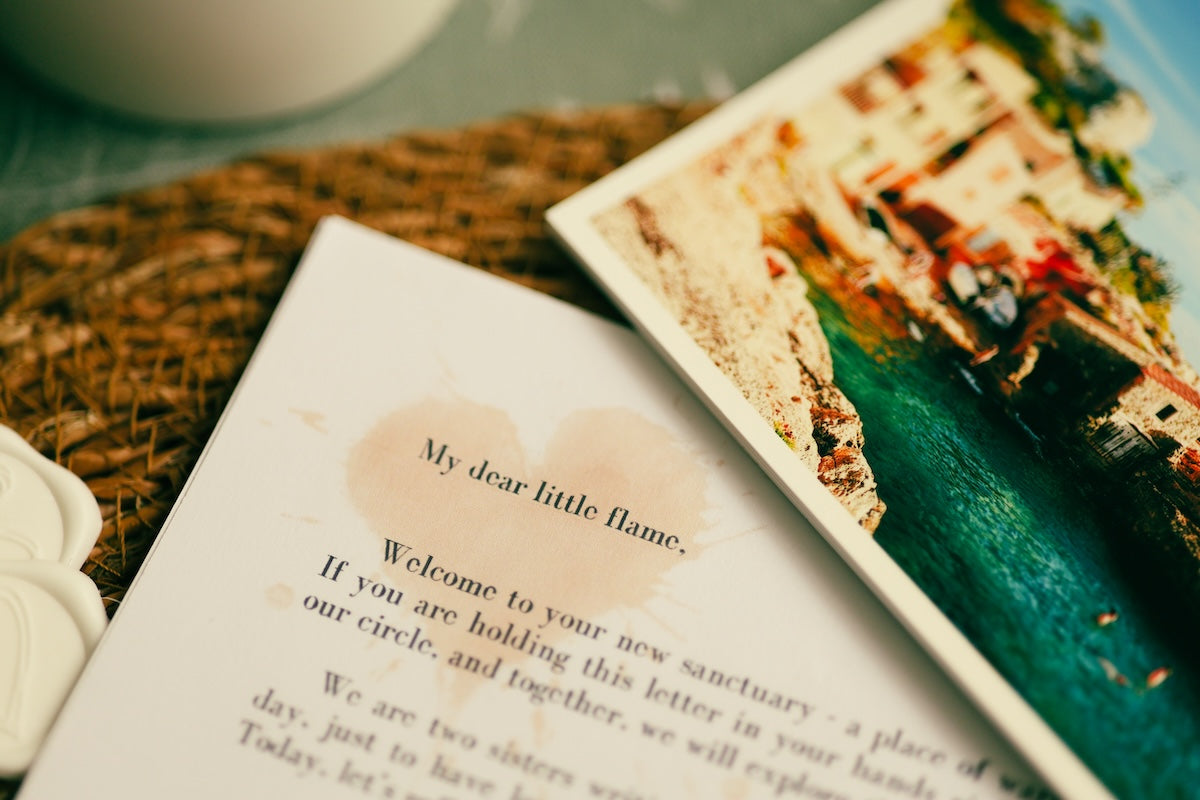 A close-up of a printed card or letter with a pastel pink watercolor wash and the text 'My dear little flame,' placed beside a printed scenic postcard showing a coastal town and turquoise water. Close up of a printed narrative letter starting with the words My dear little flame, next to a scenic travel photo print.