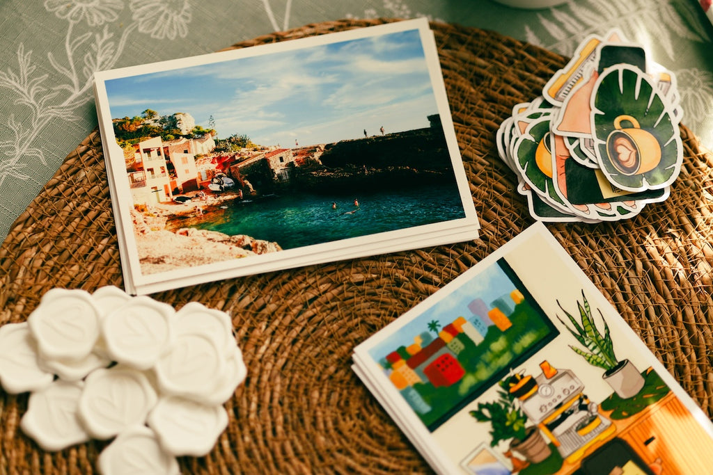 A styled flat-lay of printed cards and small white hexagonal tiles on a woven brown mat. One card shows a coastal harbor scene, another has colorful abstract artwork, and a stack of die-cut stickers is visible on the right. A scenic beach postcard, illustrated art print, a pile of white heart wax seals, and custom stickers neatly arranged on a woven mat