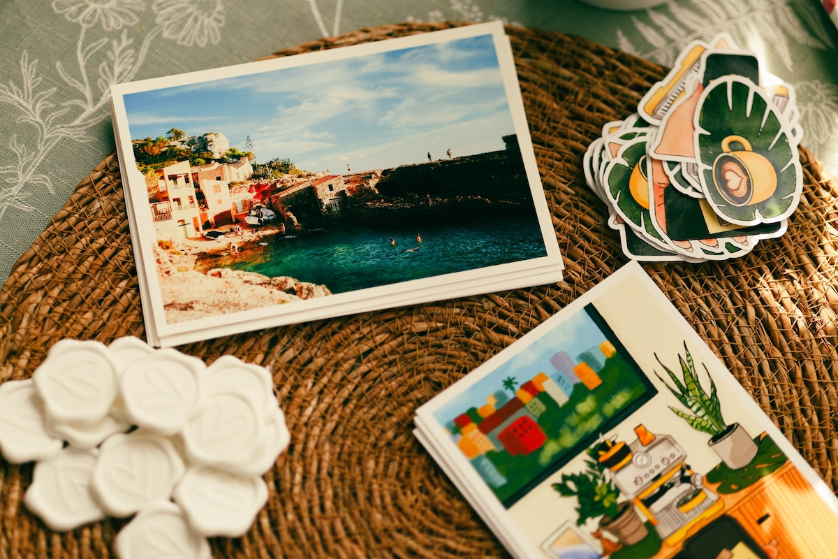 A styled flat-lay of printed cards and small white hexagonal tiles on a woven brown mat. One card shows a coastal harbor scene, another has colorful abstract artwork, and a stack of die-cut stickers is visible on the right. A scenic beach postcard, illustrated art print, a pile of white heart wax seals, and custom stickers neatly arranged on a woven mat