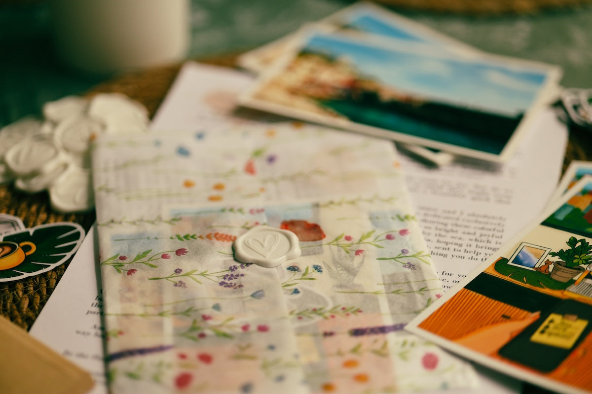 A close-up of a handmade quilted fabric envelope or letter box with a floral patchwork pattern, a white wax seal, and a ribbon closure, surrounded by scattered postcards and papers on a wooden surface. Macro shot of a white heart-shaped wax seal on a floral pattern translucent vellum envelope wrapper