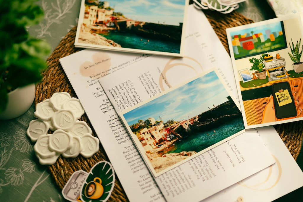 A styled flat-lay of printed postcards or photo prints with coastal and interior scenes, arranged on top of open book pages with text, alongside ceramic flowers, a small plant, and decorative paper on a woven mat. A physical travel photography print of a seaside coastal village placed over a monthly storytelling letter